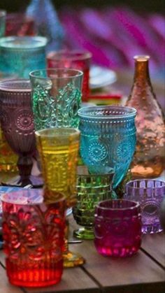 many different colored glasses are sitting on a wooden table with plates and utensils