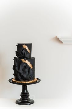 a three tiered cake with black icing and gold decorations on top, sitting on a pedestal