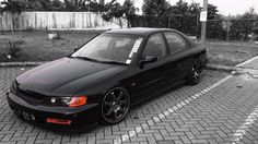 a black car parked in a parking lot next to a brick walkway and fenced in area