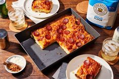 two slices of pepperoni pizza in a pan on top of a wooden table with plates and cups