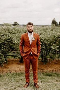 a man wearing an orange suit and tie standing in front of some bushes with his hands in his pockets