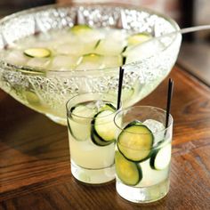 two glasses filled with cucumbers sitting on top of a wooden table next to a bowl