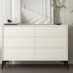 a white dresser sitting next to a plant and a mirror on top of the dresser