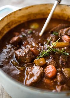 a pot filled with beef and carrots on top of a wooden table next to a spoon