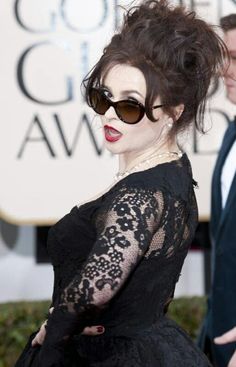 a woman in black dress and sunglasses on red carpet with man in suit behind her