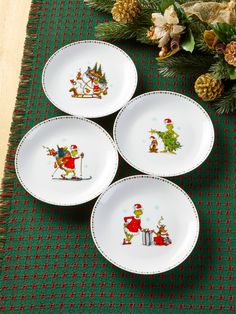 four plates with christmas designs on them sitting on a green tablecloth next to pine cones