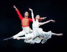 two people in white dresses and red jackets are performing on stage with one person holding the other's leg