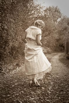 a woman in a white dress walking down a dirt road with trees and bushes behind her