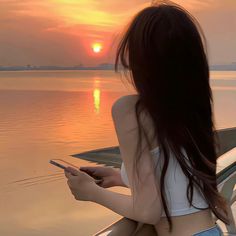 a woman sitting in a boat looking at her cell phone as the sun sets over water