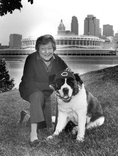 a woman kneeling next to a dog on top of a grass covered field in front of a city skyline