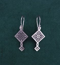 two silver earrings with designs on them sitting on a green surface, next to each other