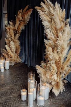 some white candles are sitting on a table with tall pamodia plants in front of them