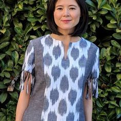 a woman standing in front of a green bush wearing a gray and white top with tassels