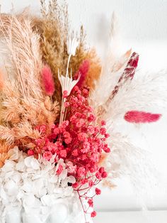 an arrangement of dried flowers and feathers in a white vase on a countertop,