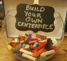 a bowl filled with legos sitting on top of a wooden table next to a sign that says build your own centerpiece