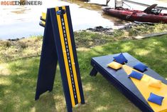 two cornhole game boards sitting in the grass next to a body of water and canoes