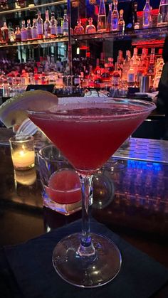 a close up of a drink in a glass on a table with liquor bottles behind it