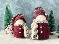 two knitted snowmen standing next to each other in front of small christmas trees