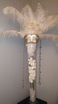 a tall vase with white flowers and feathers in it sitting on a table next to a wall