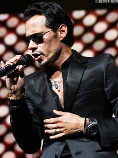 a man in a suit and sunglasses holding a microphone up to his chest while standing on stage