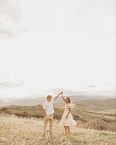 a man and woman holding hands while standing on top of a grass covered hill with mountains in the background