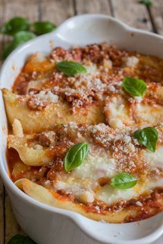 a white casserole dish filled with pasta and sauce on a wooden table next to fresh basil leaves