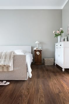 a white bed sitting in a bedroom next to a dresser and lamp on top of a hard wood floor