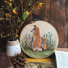 a cross stitched picture of a fox and her cub sitting on a table next to some flowers