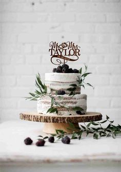 a three tiered cake with berries and greenery on top, topped with a monogram