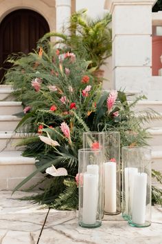 three tall vases filled with white candles sitting on top of a stone floor next to plants