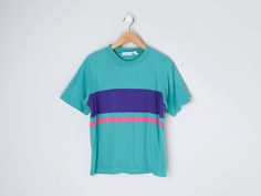 This vintage 90s t-shirt has one pink & one purple stripe. The tag size is L - it measures 21" across the chest laid flat, and it measures 24" long shoulder-to-hem. This item is in excellent vintage condition.   Content + Care - 100% cotton - Machine wash warm. Tumble dry low. Size + Fit - Tag size is L   - Chest: 21" across    - Length: 24" long shoulder-to-hem 90s Style Green T-shirt For Spring, Vintage Purple T-shirt For Summer, 90s Striped Crew Neck Top, Striped 90s Style T-shirt For Summer, Striped 90s Style Summer T-shirt, 90s Striped T-shirt For Summer, Green Short Sleeve T-shirt With Three Stripes, 90s Striped Short Sleeve T-shirt, 90s Striped Short Sleeve Tops