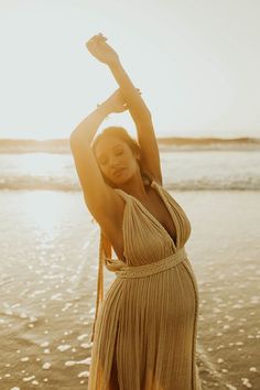 a woman in a tan dress standing on the beach with her arms above her head