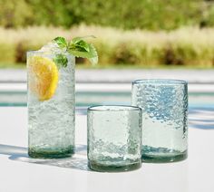 three glasses filled with water, lemon and mint sitting on a table next to a pool