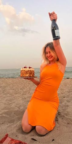 a woman in an orange dress holding up a cake on the beach with a bottle of wine