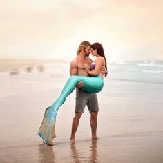 a man holding a mermaid tail while kissing a woman on the beach in front of him