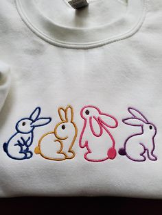 Let your cuteness factor soar with this sweatshirt featuring four embroidered bunnies. Perfect for any gender, this cozy top is adorned with adorable bunny outlines, adding a playful touch to your wardrobe. Stay warm and charming with this unique piece. (Elevate your wardrobe with some bunny love!) PRODUCT DETAILS ✦ Sweatshirts are Unisex Gildan Heavy Blend G180 ✦ 50% USA Cotton, 50% Polyester 13.3 oz./lin.yd. ✦ Pill-resistant air jet yarn ✦ Double needle stitching throughout ✦ Set in sleeves ✦ Cute White Embroidered Sweatshirt, Embroidered Bunnies, Bunny Sweatshirt, Adorable Bunny, Cozy Tops, Bunny Shirt, Embroidered Shirt, Air Jet, Cute Bunny