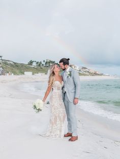 a bride and groom kissing on the beach with a rainbow in the sky behind them