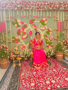 a woman in a pink sari sitting on a red rug with flowers and greenery behind her