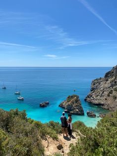 two people standing on a cliff looking out at the ocean and boats in the water