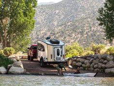 two people are setting up their camper on the side of a river while another person stands next to it