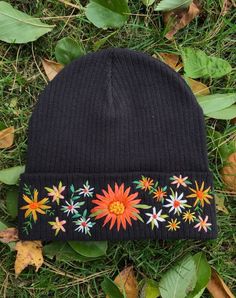 a black beanie with colorful flowers on it laying in the grass next to leaves