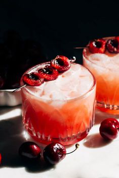 two glasses filled with ice and cherries next to some cherries on the table