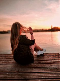 a woman sitting on a dock with her hair blowing in the wind and drinking water