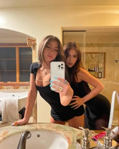 two beautiful young women standing next to each other in a kitchen looking at a cell phone