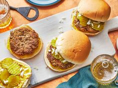 three hamburgers with pickles on a cutting board next to two glasses of beer