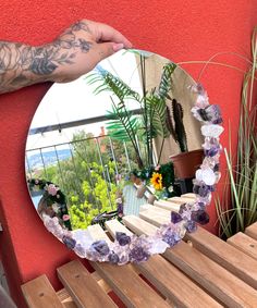 a person holding up a mirror with flowers on it and plants in the back ground