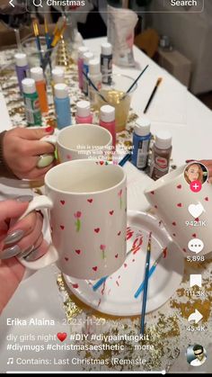 two people are holding cups on a table with paintbrushes and other items around them