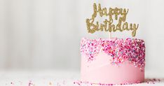 a pink birthday cake with sprinkles and a gold happy birthday topper