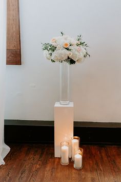 white flowers in a tall glass vase with candles on the floor next to each other