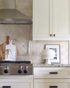 An off-white subway tile backsplash is installed next to a decorative off-white herringbone tile backsplash. A stainless steel hood hangs above the stainless steel range. A gray counter is laid on the white cabinets...   Image: lavenderrosehome